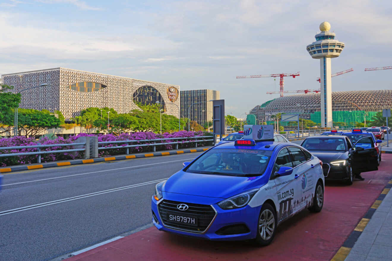 Blue taxis lined up on a road near a futuristic airport control tower and modern buildings, with cranes in the background.