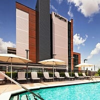 Outdoor pool with lounge chairs and umbrellas in front of a modern multi-story hotel building with “Westin” signage against a partly cloudy sky.
