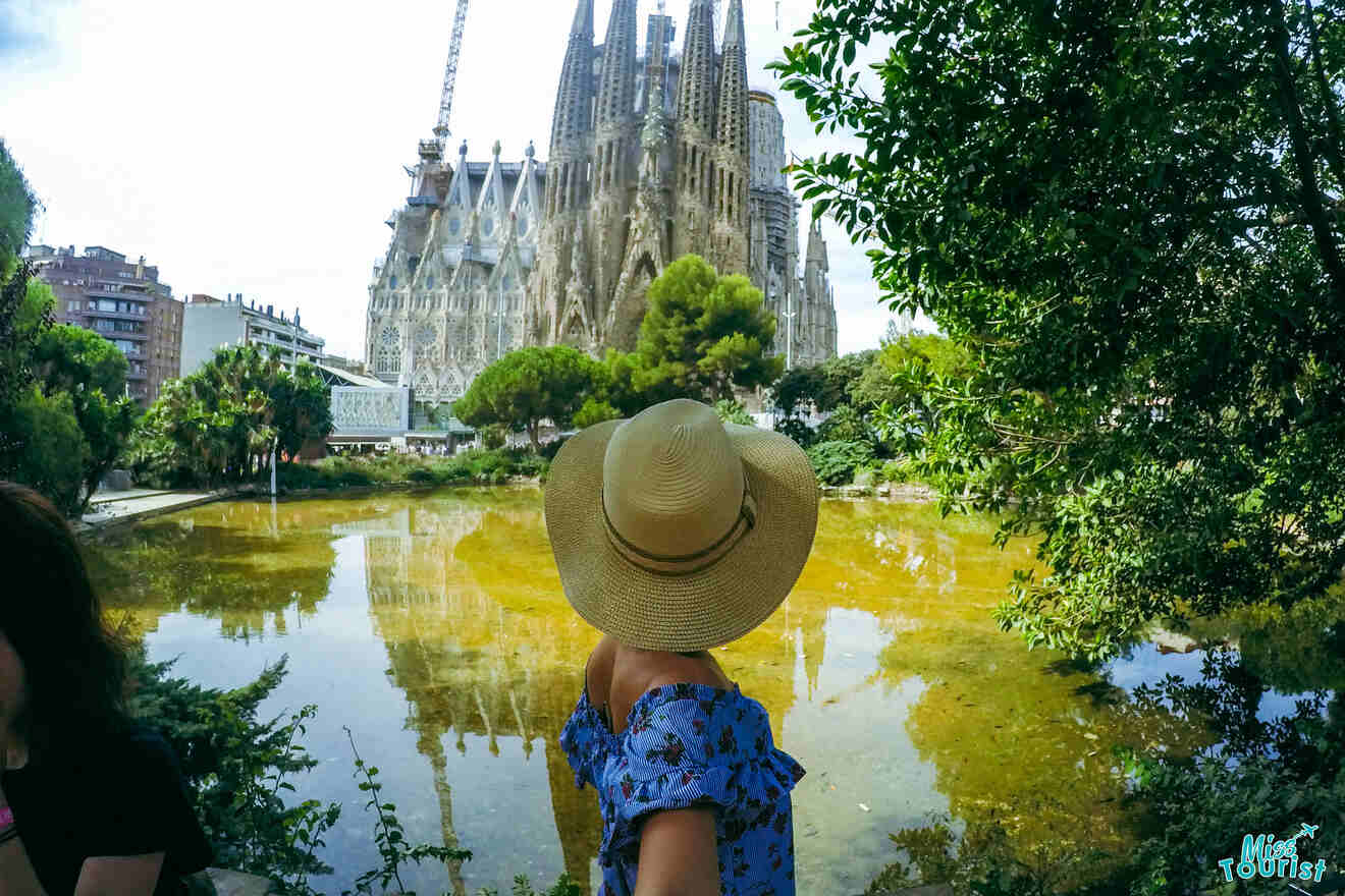 author of the post with a wide-brimmed hat and blue dress stands in front of a pond, with the Sagrada Familia basilica in Barcelona visible in the background.