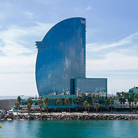 Tall, modern, sail-shaped glass building with a "W" on top, located by the waterfront with palm trees and a clear blue sky.