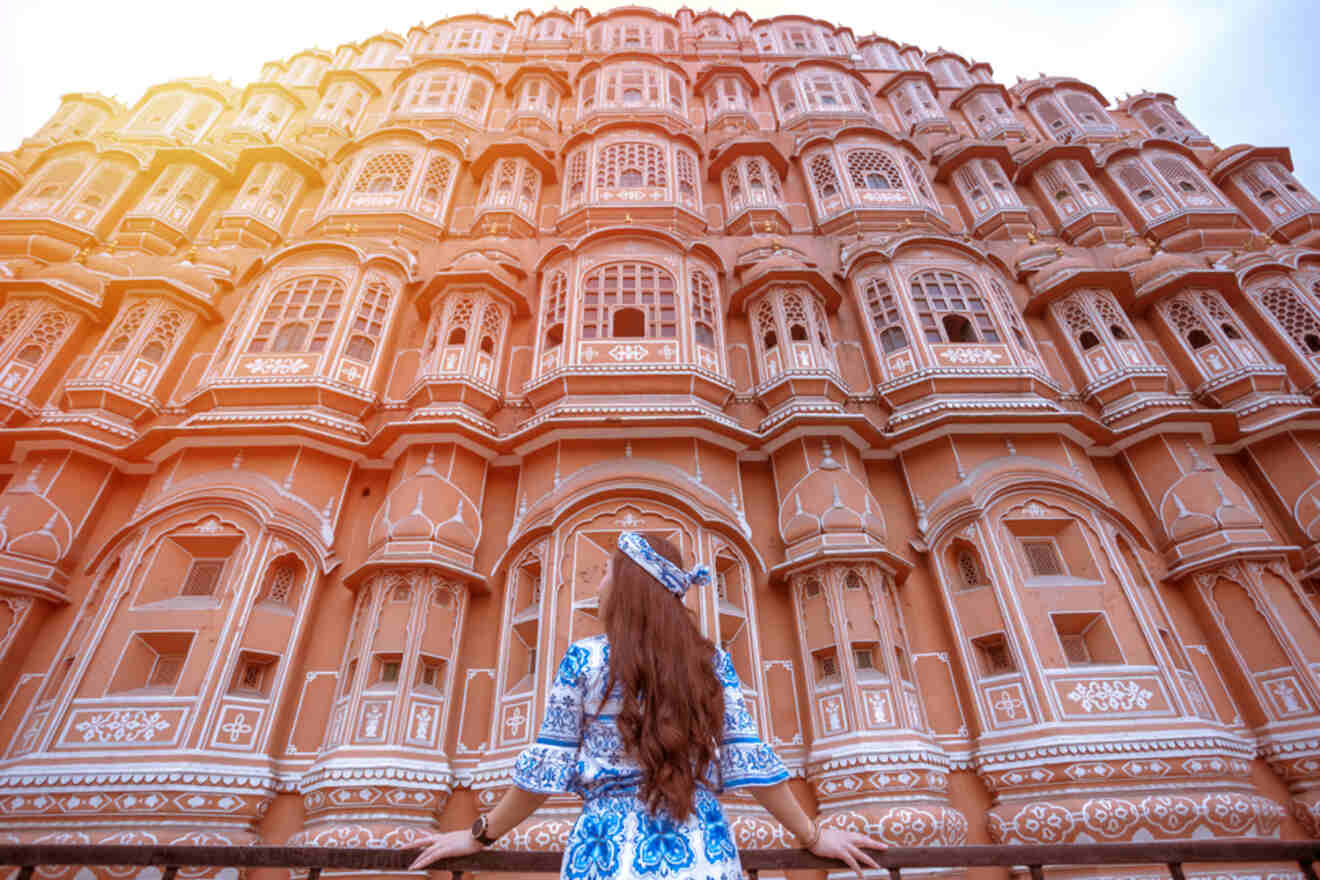 A person wearing a blue and white outfit faces an elaborate building with numerous windows and intricate designs, seen from behind. The building is bathed in warm sunlight.