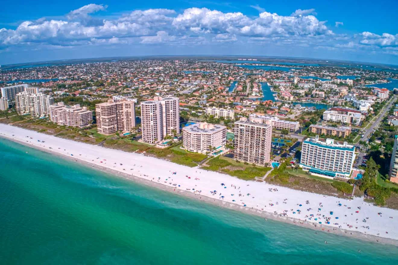 Aerial view of a coastal city with high-rise buildings along a sandy beach and turquoise waters. The cityscape features numerous residential areas, roads, and waterways under a partly cloudy sky.