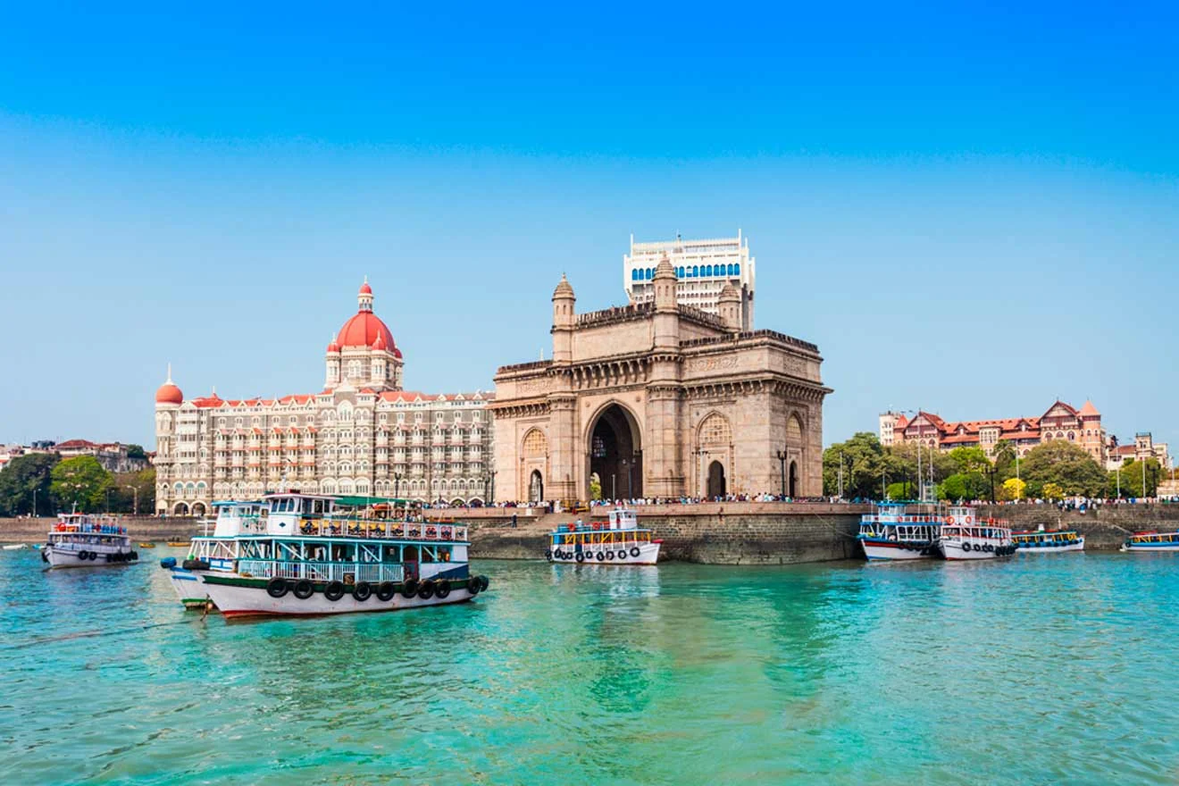 Several boats float on the water in front of the Gateway of India and Taj Mahal Palace Hotel under a clear blue sky.