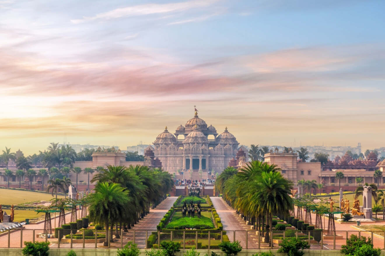 A wide view of the Akshardham Temple in Delhi, India, surrounded by manicured gardens and palm trees, under a colorful sunset sky.