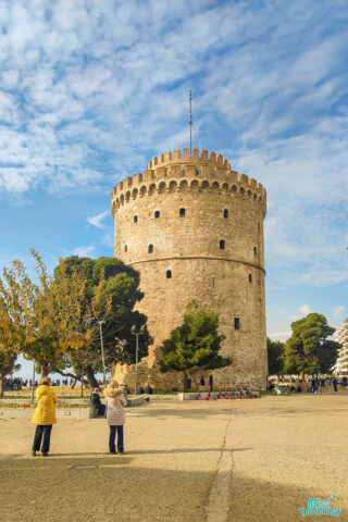 The image shows the White Tower of Thessaloniki, a cylindrical structure with a crenellated top, situated on a stone-paved area with people walking and trees surrounding the scene.