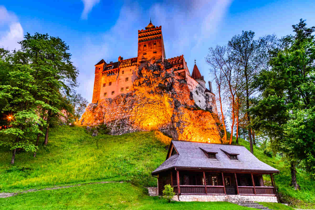 A historic castle with illuminated walls stands atop a rocky hill, surrounded by lush trees and a small wooden cabin at the base, under a blue sky.