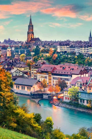 Aerial view of a picturesque city with colorful buildings, a church with a tall spire, and a river winding through it during sunset.