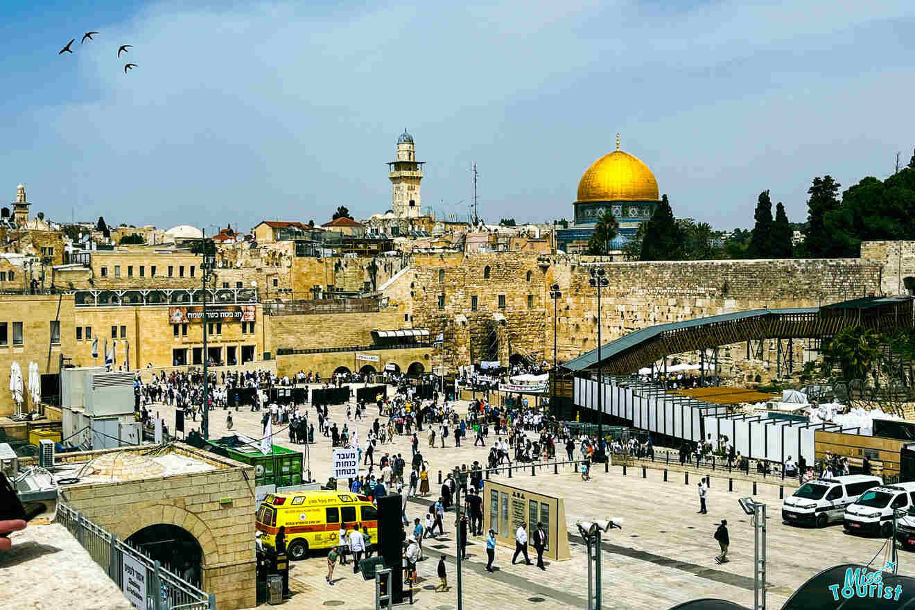 A large courtyard filled with people is shown with historic buildings in the background, including a mosque with a golden dome and the Western Wall. An ambulance is parked at the side. Birds fly in the sky.