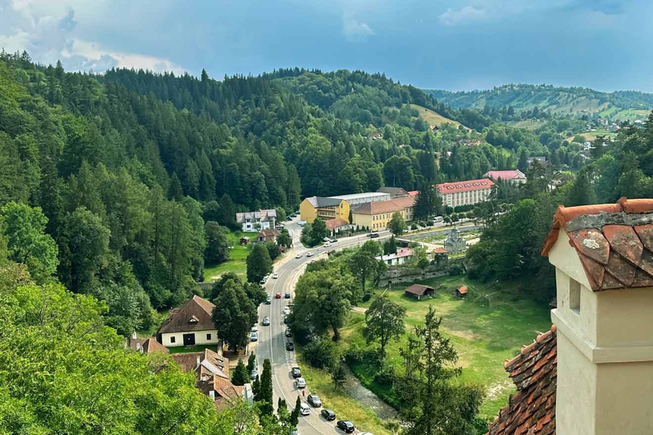 A winding road passes through a green, hilly landscape with scattered buildings and dense forests in the background, under a slightly overcast sky.