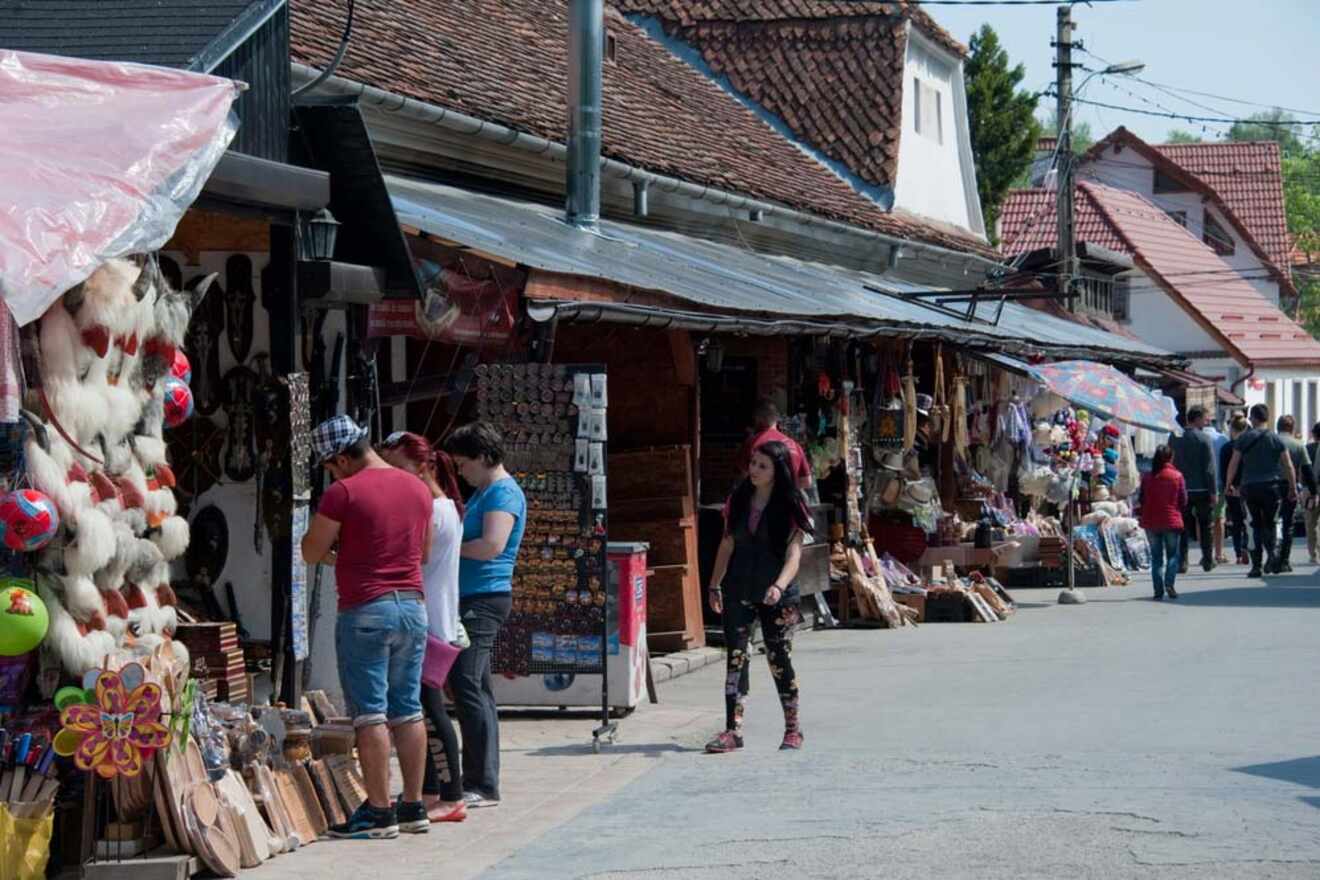 People browsing various stalls at an open-air market, featuring handmade crafts, decorations, and souvenirs. Red-roofed buildings line the street.