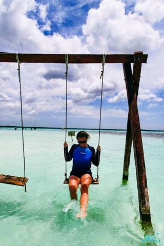 Person wearing a blue rash guard sitting on a swing set in shallow, clear ocean water under a partly cloudy sky.