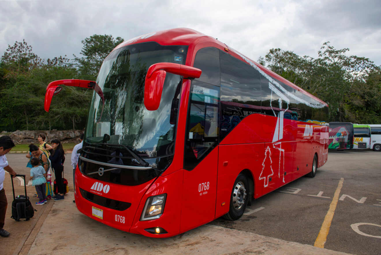 A red ADO bus with the number 0768 is parked at a bus terminal under a cloudy sky. Passengers with luggage are seen near the bus.