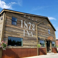 Wooden building with "1872 INN" painted on it, featuring two windows and hanging flower baskets, under a blue sky.