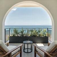 Ocean view from a balcony with two cushioned wooden chairs, a small round table, and a railing with plants under a curved archway.