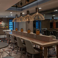 Modern restaurant interior with long communal table, pendant lights, and upholstered chairs. Bar area with illuminated shelves visible in the background.