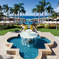 A resort pool area with a fountain in the foreground, surrounded by palm trees and cabanas, overlooking the ocean.