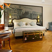 Hotel room with a large bed, vintage-style trunk at the foot, mural on the wall, wooden floor, and a table with a plant and coffee cup.