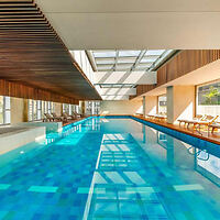 Indoor swimming pool with clear water, surrounded by lounge chairs and wooden accents under a skylight ceiling.