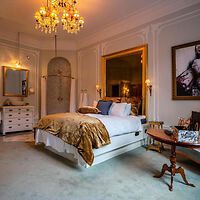 Elegant bedroom with a large bed, gold bedding, chandelier, wall sconces, and framed art. A round wooden table and white dresser are present. Classic decor and warm lighting create a cozy ambiance.