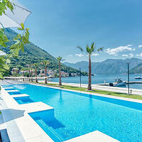 A pristine swimming pool extends towards a scenic view of mountains and a lake, bordered by palm trees and lined with sun loungers.