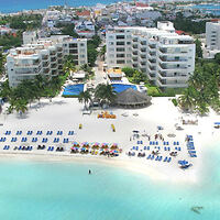 Aerial view of a beach resort with several white buildings, a pool, rows of beach chairs, umbrellas, and a clear blue sea in the foreground.