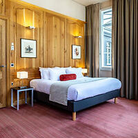 Hotel room with a large bed, red bolster pillow, wooden paneling on the wall, two lamps, framed artwork, and a window with long curtains.