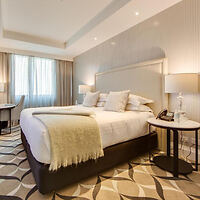 Hotel room with a large bed, pillows, and a beige throw. Two bedside tables with lamps, and a desk with a chair near a window with curtains. Patterned carpet covers the floor.