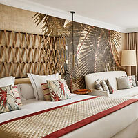 A hotel room with a double bed, patterned pillows, a decorative headboard, and a textured wall mural featuring palm leaves.