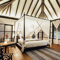 A cozy bedroom with a four-poster bed draped in white fabric. The room features wooden floors, a high ceiling with exposed beams, and a small seating area with a table and chairs.