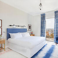 Bright bedroom with a king-sized bed, blue headboard, patterned curtains, and a balcony view. Decor includes a rug, bedside tables with lamps, and framed art on the walls.