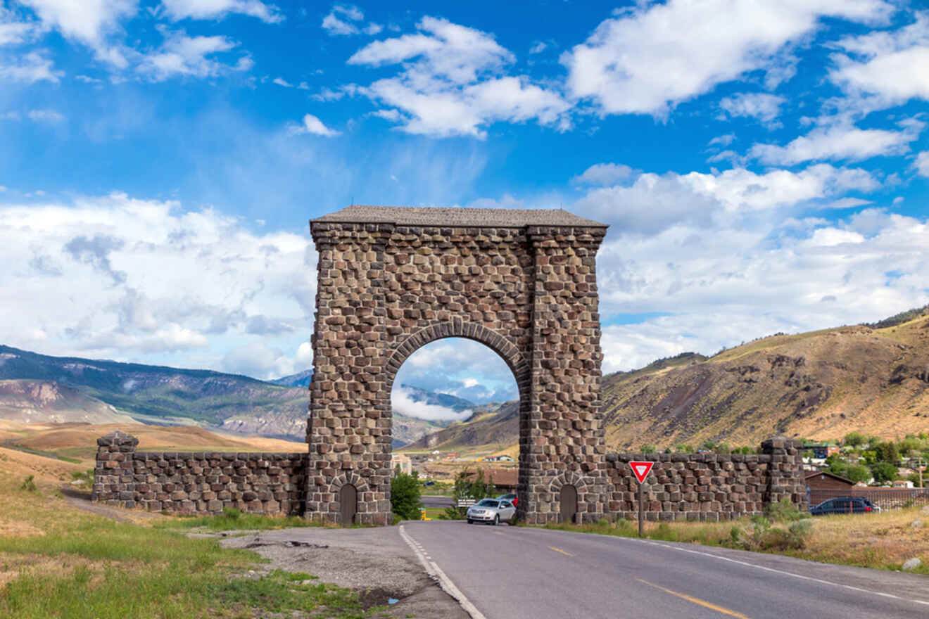 A stone archway stands over a road in a rural landscape, with hills and clouds in the background. A sign indicates a yield.