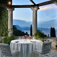 A round table with a white tablecloth, set for two with flowers, stands on a balcony overlooking a scenic view of the sea and distant mountains, surrounded by columns and greenery.