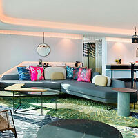 Modern living room with a curved gray sofa, colorful cushions, round mirror on the wall, and abstract-patterned rug. Hanging lights and a view of the kitchen in the background.