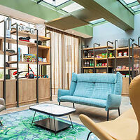 A modern lounge with a blue sofa, chairs, shelves with various items, and a glass ceiling, featuring wooden accents and a green rug.