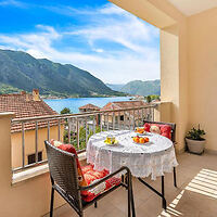 A balcony with two chairs and a table set for breakfast overlooking a coastal view with mountains and rooftops.