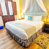 A neatly made bed with a patterned headboard and light bedding in a brightly lit room with wood flooring. Two bedside tables with lamps are on either side, and a patterned rug lies beside the bed.