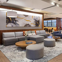 Modern lounge area with a large gray sectional sofa, round wooden tables, gray ottomans, and abstract artwork on the wall. Soft lighting from ceiling fixtures and ample natural light from windows.