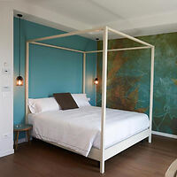 A bedroom with a four-poster bed, white bedding, teal accent wall, and wooden floor. Two hanging pendant lights are on either side of the bed.