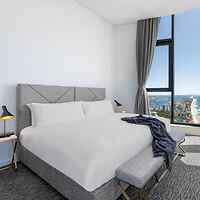 Modern bedroom with a large bed, gray headboard, two bedside lamps, an upholstered bench, and floor-to-ceiling windows offering a city and ocean view. A gray blanket is draped over the bed.