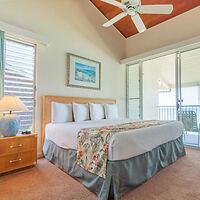 A bedroom with a double bed, floral blanket, two pillows, a bedside table with a lamp, a ceiling fan, and sliding glass doors leading to a balcony.