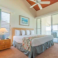 A bedroom with a double bed, floral blanket, two pillows, a bedside table with a lamp, a ceiling fan, and sliding glass doors leading to a balcony.