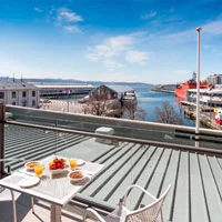 A balcony overlooking a harbor with tables set for breakfast. Two plates with food, two glasses of orange juice, and a bowl of fruit are on the table. A scenic view of water and boats is visible.