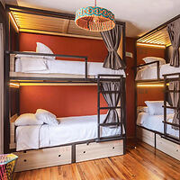 A room with two sets of bunk beds, each with curtains for privacy, placed against an orange wall. There is a wooden floor and a light fixture above.