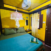 A cozy bedroom with yellow walls, a double bed with green sheets, a small side table, and a ceiling fan. A towel is neatly folded on the bed.