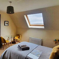 A bedroom with a skylight, double bed, and two folded towels. Two yellow chairs and a small glass table are in the corner. A pendant lamp hangs from the ceiling.