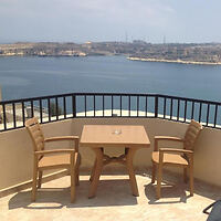 A balcony with a wooden table and two chairs overlooks a body of water with distant land on the horizon.