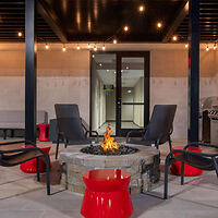 Outdoor patio scene with four black chairs around a lit fire pit. Red stools are nearby, and string lights hang above. Glass doors are in the background.