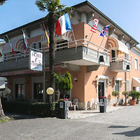 Two-story hotel with various international flags displayed. Outdoor seating is visible near the entrance, and a tree stands in front of the building.