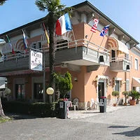 Two-story hotel with various international flags displayed. Outdoor seating is visible near the entrance, and a tree stands in front of the building.