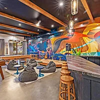 Colorful room with a mural, featuring seating options including stools, bean bags, and small tables, under wooden beams and pendant lights.